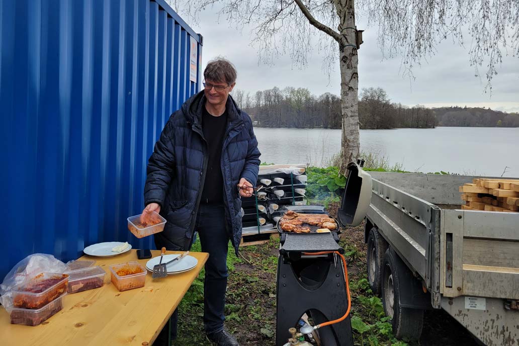 Volker Gawron steht an einem Grill auf der Baustelle der neuen Tribüne an den Eutiner Festspielen. Im Hintergund der Eutiner See hinter einer Birke.