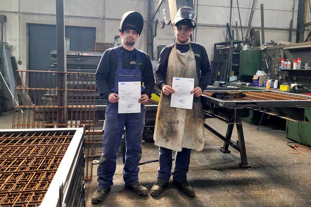 Zwei Metallbaulehrlinge in der Werkstatt halten die Zeugnisse eines Schweißlehrgangs in den Händen.