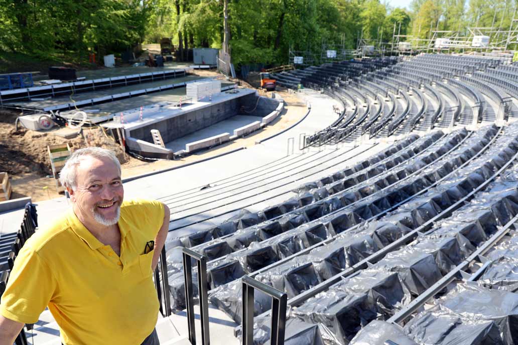 Ein Mann in gelbem T-Shirt lacht links unten im Bild in die Kamera. Rechts im Bild die Baustelle der Eutiner Festspiele.