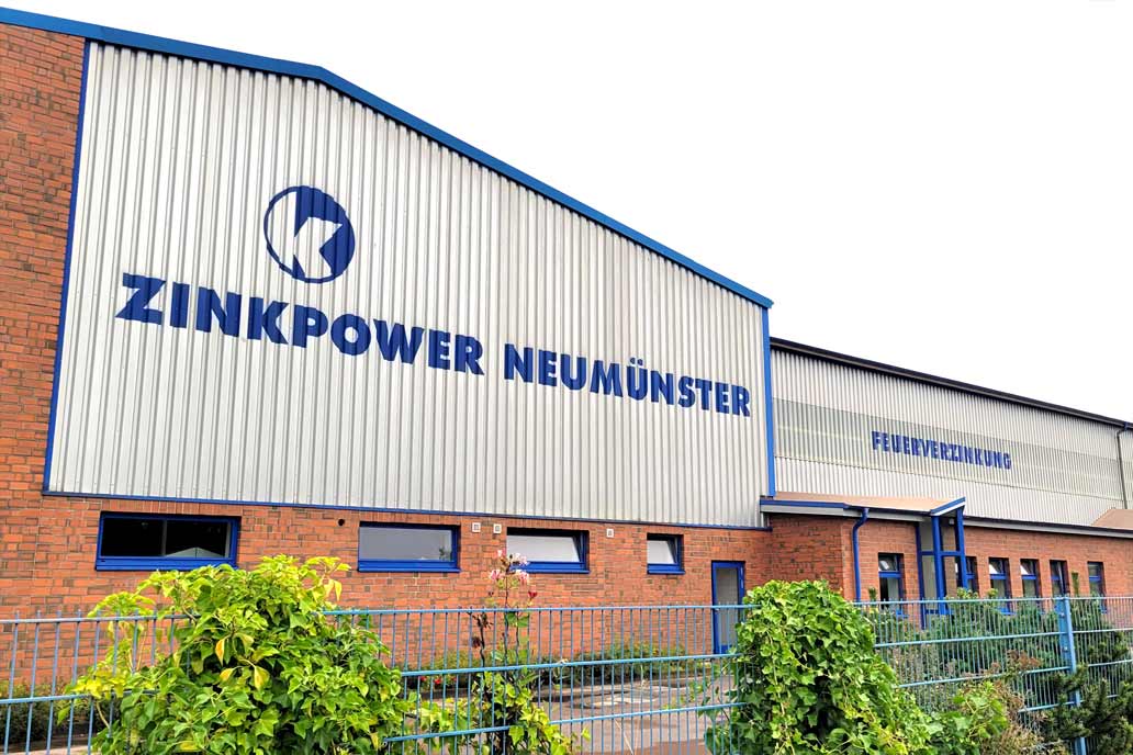 Fassade einer Industriehalle mit dem blauen Schriftzug "ZINKPOWER NEUMÜNSER" und "FEUERVERZINKUNG".