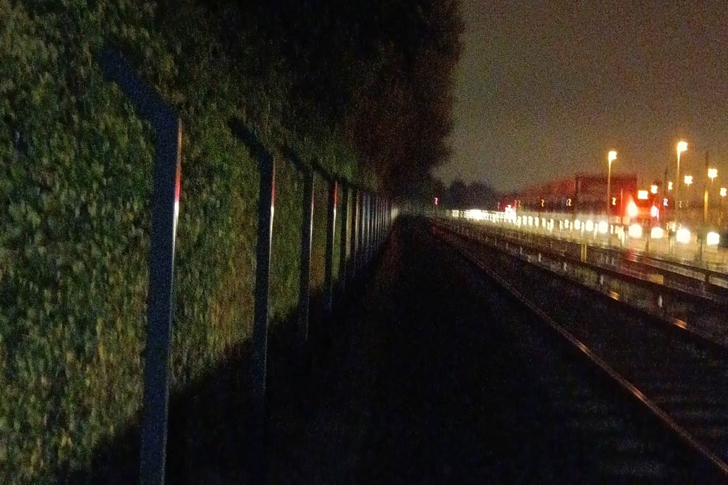Ein unscharfes Foto von Gleisen. Links neu gesetzte Zaunpfosten, rechts im Bild viele helle Lampen entlang eines Bahnsteigs.