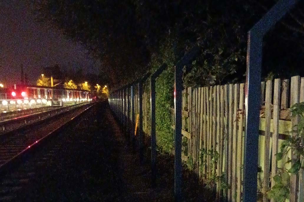 Gleisanlage im Dunkeln. Rechts neu gesetzte Zaunpfosten, links viele helle Lampen entlang eines Bahnsteigs.