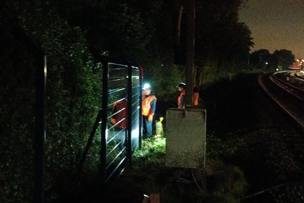 Mehrere Arbeiter in orangenen Warnwesten mit Kopflampen während der Zaun Montage in dunkler Nacht an den S-Bahn Gleisen.