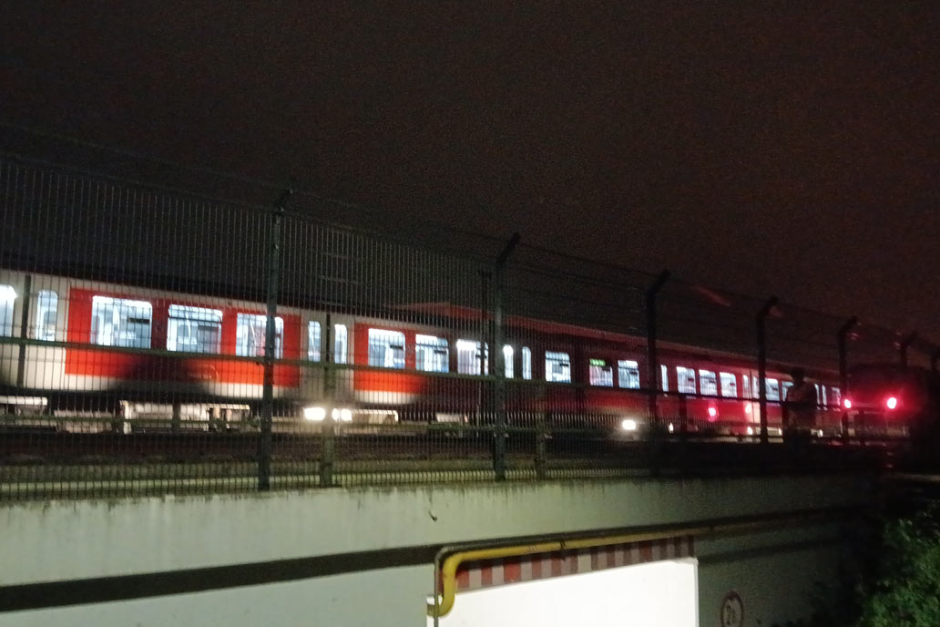 Ein hell erleuchteter rot-weißer Zug der S-Bah Hamburg im Dunkeln hinter einem neu montierten Stahlmattenzaun.