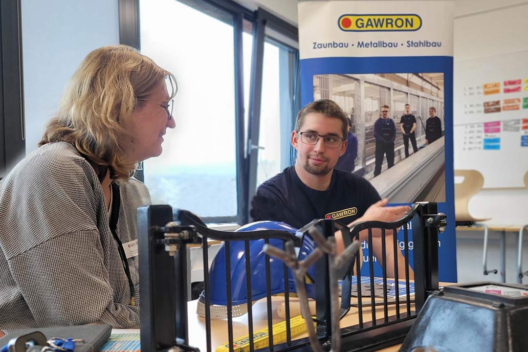 Eine Frau und ein Mann unterhalten sich auf einer Berufsmesse. Im Vordergrund ein Miniatur Metalltor, im Hintergrund ein Rollup von Gawron mit der Aufschrift: Zaunbau - Metallbau - Stahlbau.