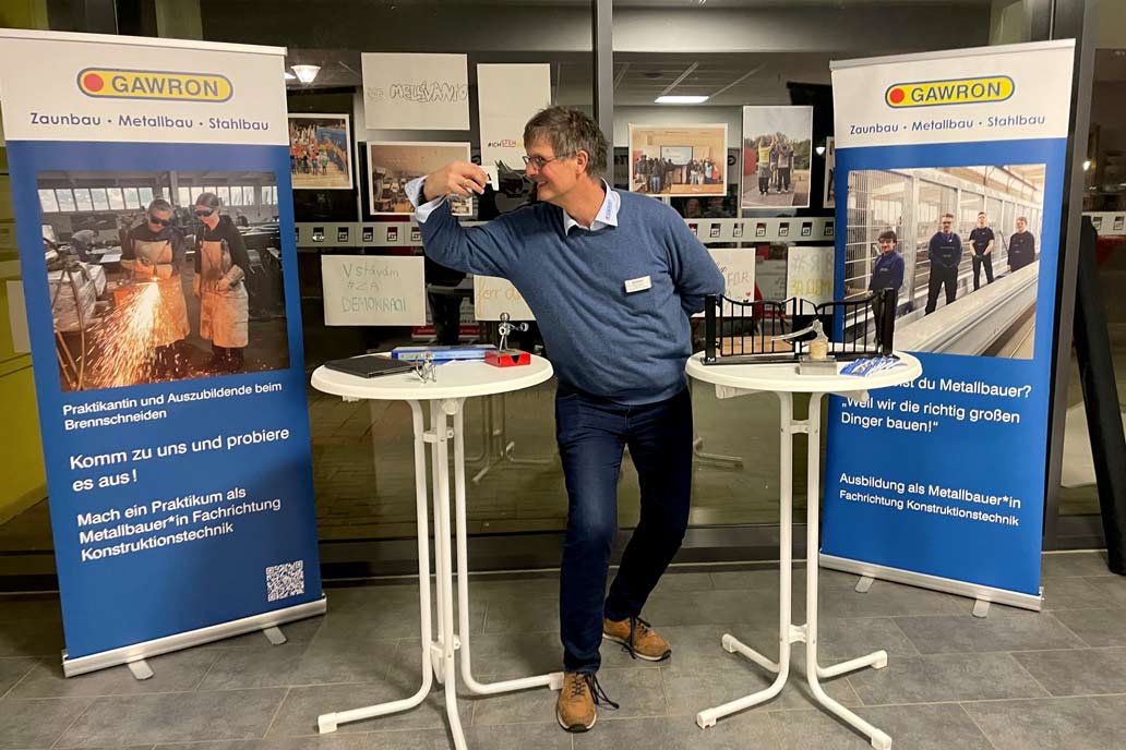 Volker Gawron steht zwischen zwei Stehtischen am Infostand auf dem Infoabend Berufliche Orientierung in Halstenbek an der Gemeinschaftsschule an der Bek. Er sieht dabei einem Elefanten aus Blech in die Augen. Im Hintergrund zwei blaue Rollups mit Metallbau Azubis.
