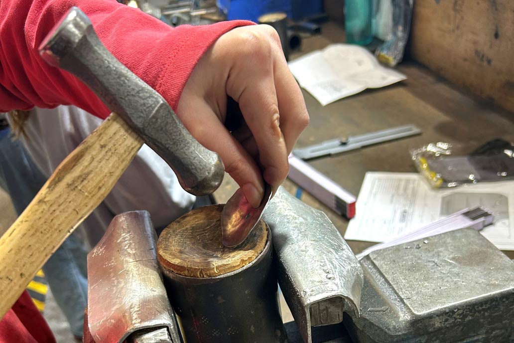 Die Hand eines Mädchens im Bildausschnitt: Sie dengelt ein Blech mit einem Hammer im Metallbaubetrieb bei Gawron & Co.