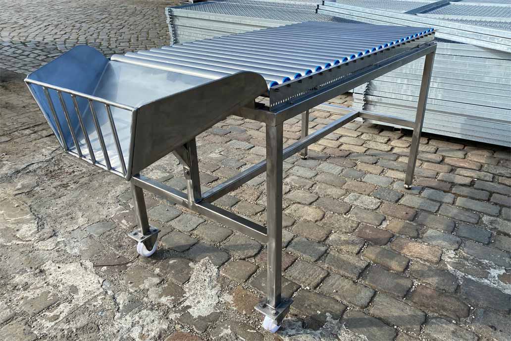 Ein Arbeitstisch mit Rollenband und einem schrägen Auslauf aus Edelstahl zum Abfüllen von Ware in Säcken in weiterverarbeitende Maschinen steht auf dem gepflasterten Hof vom Metallbaubetrieb Gawron in Rellingen.