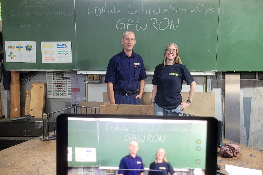 Technik-Check für eine Online Veranstaltung: Metallbaumeister und Ausbilder Kai Potthoff und Büromanagement Azubi Katharina stehen in der Lehrwerkstatt vor einer grünen Tafel auf der steht: Digitale Lehrstellenrallye – Gawron. Unten im Bild die unscharfe Abbildung desselben Motivs auf einem Tablet.