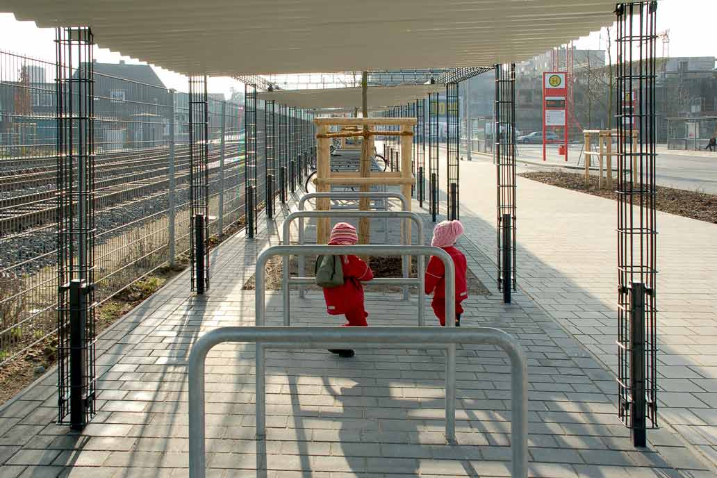 Zwei kleine rot gekleidete Kinder spielen an gebogenen Fahrradbügeln aus feuerverzinktem Stahl unter einem wellblachdach das von Rankpfosten getragen wird. Im Hintergund der Bahnhof Quickborn.
