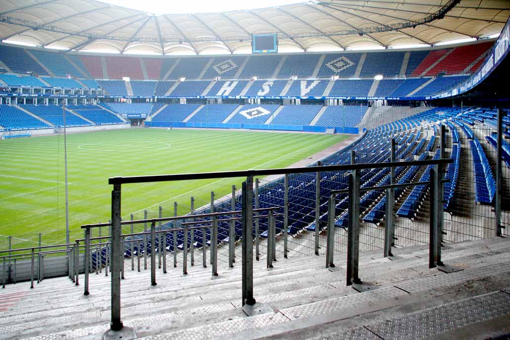 Foto mit Blick ins Volksparkstadion Hamburg. Im Vordergrund Wellenbrecher an den Stehplätzen, rechts ein Doppelstabzaun vor den Sitzplätzen.