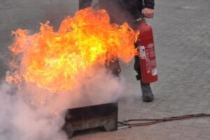 Eine orange lodernde Flamme, dahinter ein Mann mit einem Feuerlöscher.