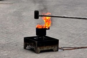 Ein Metallbehälter an einer langen Stange mit Wasser über einem Stahlbehälter mit einer niedrigen orangenen Flamme brennenden Öls.