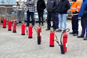 Sieben rote Feuerlöscher stehen in Reihe vor einer Gruppe Menschen auf dem gepflasterten Hof von Gawron in Rellingen.