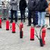Sieben rote Feuerlöscher stehen in Reihe vor einer Gruppe Menschen auf dem gepflasterten Hof von Gawron in Rellingen bei der Brandschutzschulung.
