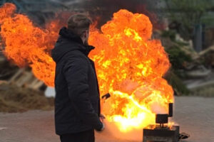 Eine orange-gelbe Wolke bei einer Fettbrandexplosion im Rahmen der Brandschutzschulung beim Metallbauunternehmen Gawron. Im Vordergrund steht ein dunkel gekleideter Mann, der kurz zuvor an einer langen Stange etwas Wasser in brennendes Öl geschüttet hatte.