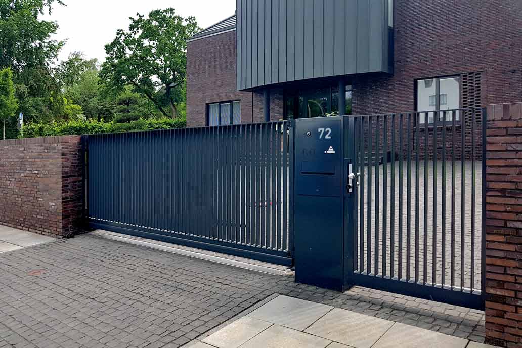 Ein dunkelgraues Schiebetor mit Flachstabfüllung mit einer Personentür im selben Design rechts daneben. Eingepasst in eine Mauer aus dunklem Rotklinker. Rechts dahinter ein modernes Haus in demselben dunklen Klinker, lnks hohe grüne Bäume.